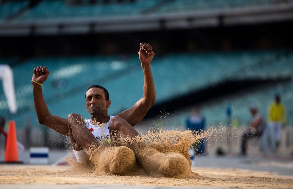 Dynamic action shot of a male athlete executing a long jump in a stadium setting.