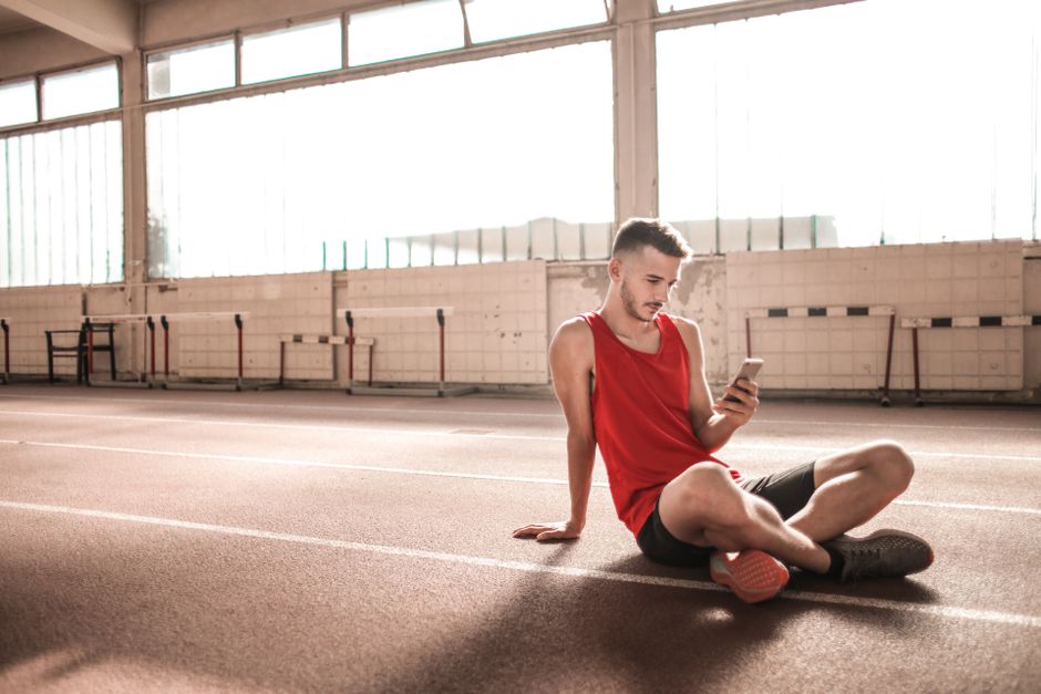 Young male athlete in red sportswear sitting on indoor track using smartphone, embracing modern technology
