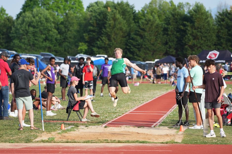 Male athlete performing a long jump at a sunny outdoor track and field meet with spectators.